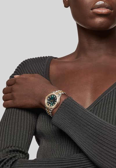 Guld- och silverklocka med en rund grön urtavla, diamantdetaljer och ett länkarmband. Bärs på en mörk, texturerad och räfflad topp.