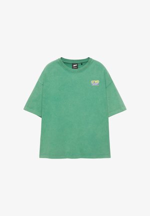 Jeune homme debout contre un mur blanc, portant un t-shirt vert, un short cargo marron foncé, des chaussettes blanches, des baskets grises et un collier de perles.