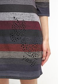 Robe rayée dans des nuances de gris, bordeaux et marine avec un design floral découpé au laser en bas à gauche. Tissu doux et texturé.