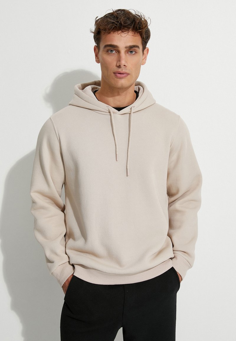 Sudadera beige con un bolsillo frontal tipo pouch, capucha con cordón y puños acanalados. Textura suave; combinada con pantalones oscuros.