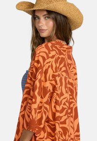 Kimono con patrón naranja y diseño fluido que presenta motivos de hojas. Llevado sobre una blusa a rayas azul, complementado con un sombrero de paja.