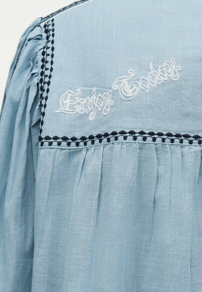 Ανοιχτό μπλε ύφασμα με μαύρο περιθώριο και άσπρο κεντημένο κείμενο «Enjoy Today» σε διακοσμητικό στυλ γραφής, με πιέτες κάτω από το κέντημα.