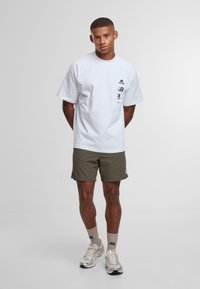 Joven de pie con las manos detrás de la espalda, vestido con una camiseta gráfica blanca, pantalones cortos verde oliva, calcetines beige y zapatillas de correr plateadas.