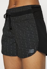 Černé sportovní šortky s mřížkovým vzorem bílých čar, elastický pas se šňůrkou a logo New Balance na spodním lemu.
