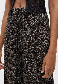Pantalon noir à motifs avec un design floral en beige. Tissu doux avec une coupe ample, doté d'un lien à la taille et de poches latérales.