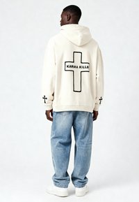 Felpa oversize color crema con una croce nera e il testo "KARMA KILLS" sulla schiena, abbinata a jeans larghi azzurri e sneaker bianche.