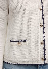 Weißer Strickcardigan mit Struktur, marineblauem Besatz, goldgeprägten Knöpfen und einer kleinen Vorderjackentasche über blauen Jeans.