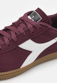 Vínové semišové tenisky s bílým bočním akcentem, texturovanými tkaničkami a gumovou podrážkou. Na jazyku je černobílá nášivka s logem.