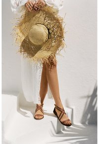 Sandali in pelle marrone con cinturini incrociati e una fibbia. Un grande cappello di paglia con bordi sfrangiati e un fiocco nero. Un cover-up bianco leggero e texturizzato.