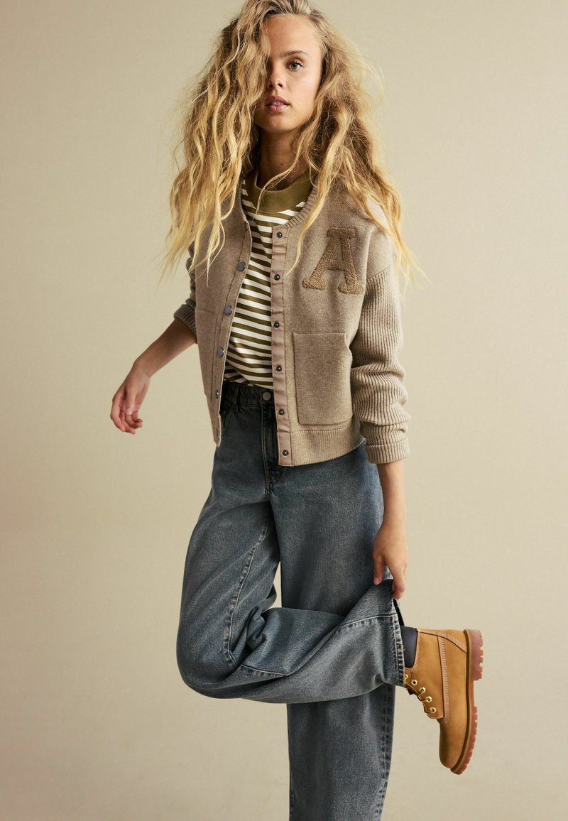 Lys brun cardigan med ribbede ærmer, prydet med et stort bogstav "A"; lagdelt over en stribet grøn og hvid skjorte samt løse denimjeans.