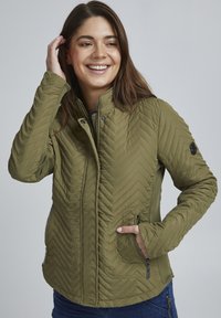 Veste matelassée vert olive avec un motif en chevrons, col haut et poches zippées. Texture douce et design ajusté.