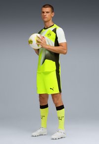 Uniforme da calcio giallo brillante con accenti neri, caratterizzato da un logo, maniche corte e pantaloni abbinati. Il giocatore tiene un pallone da calcio bianco.