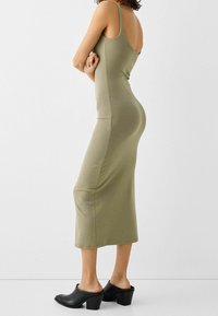 Olivově zelené, přiléhavé midi šaty s tenkými ramínky, doplněné černými podpatkovými mules. Hladký, elastický materiál, minimalistický design.