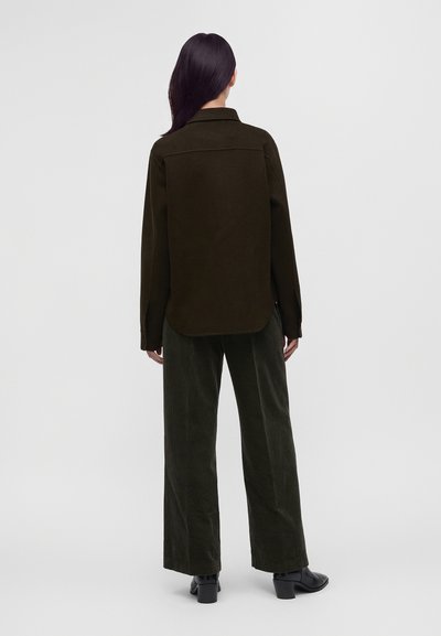 Personne aux longs cheveux foncés portant une veste vert olive, un pantalon large assorti et des bottines noires à talons, vue de dos.