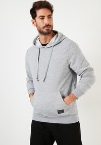 Gray cotton hoodie with a kangaroo pocket, drawstring hood, and ribbed cuffs. Features a small black logo patch on the lower front.