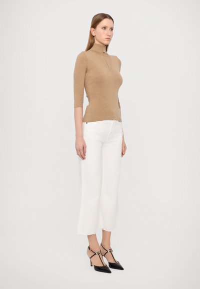 Pull en laine marron à col roulé avec des manches trois-quarts, associé à un pantalon blanc court et des escarpins noirs à bout pointu. Détails avec une fermeture éclair.