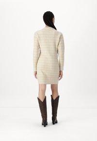 Robe beige texturée avec une silhouette ajustée, des manches longues et un col haut. Portée avec des bottes de cowboy marron jusqu'aux genoux.