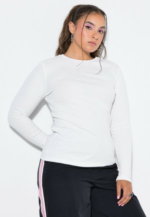 Ung kvinde med langt hår iført hvid langærmet skjorte og sorte bukser med pink striber, poserer med den ene hånd på hoften mod hvid baggrund.