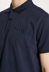 Námornícky polo tričko vyrobené z bavlny s klasickým golierom, dvoma čiernymi gombíkmi a vyšitým logom v tmavšej námorníckej farbe na hrudi.