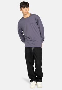 Langärmliges lila Baumwoll-T-Shirt mit einem kleinen schwarzen Logo auf der Brust, kombiniert mit lockeren schwarzen Jeans und weißen Turnschuhen.