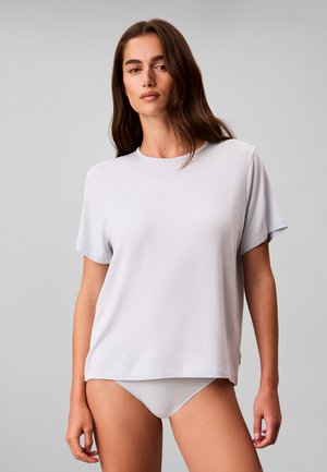 Mujer con cabello largo y castaño que lleva una camiseta lisa de manga corta color gris claro y bragas a juego, de pie frente a un fondo gris.