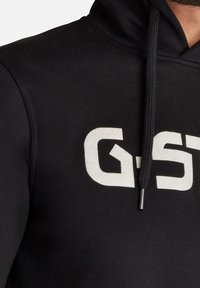 Sudadera negra con un bolsillo canguro en la parte frontal, que presenta un gran logo blanco. Fabricada con un tejido suave, cuenta con capucha con cordones y un acabado texturizado.