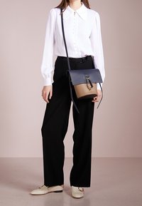 Bílé blůza s límečkem a knoflíkovou přední částí, kombinovaná s černými kalhotami se širokými nohavicemi. Drží dvoubarevnou kabelku se zlatým kováním.