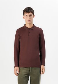 Polo shirt maro cu mânecă lungă, cu guler, trei butoane negre și un tricot texturat. Asortat cu pantaloni din catifea verde măslinie.