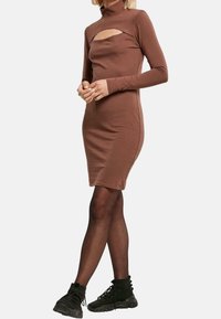 Vestito marrone a maniche lunghe con collo alto e apertura sul petto, realizzato in materiale elasticizzato, abbinato a stivaletti neri alla caviglia.