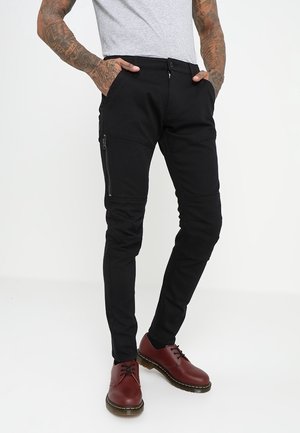 Man draagt slanke zwarte broek met zijritszakken, grijs T-shirt en donkerrode veterschoenen, staat met handen in de zakken.