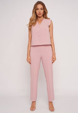 Kobieta stojąca w różowym, bezrękawnym, zapinanym na guziki topie i dopasowanych, wysokowoskich, prostych spodniach, w cielistych sandałach na obcasie, na białym tle.