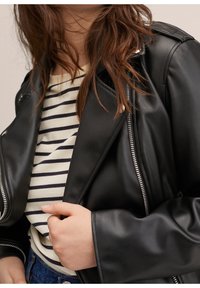 Veste noire en similicuir avec fermeture éclair argentée, poches latérales et texture lisse, associée à une chemise à rayures en dessous.