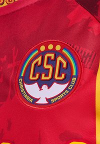 Röd sporttröja med en cirkulär patch som har en vit duva, initialerna "CSC" och texten "CHRISTIANIA SPORTS CLUB" i gult och blått.