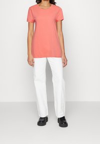 Różowa koszulka z krótkim rękawem i okrągłym dekoltem, zestawiona z białymi spodniami o prostym kroju i czarnymi butami, wyeksponowana na neutralnym tle.