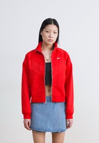 Punainen vetoketjullinen takki, jossa on korkea kaulus, pehmeästä materiaalista valmistettu, jossa on Nike-logo ja geometrisia ompelut yksityiskohtia. Yhdistetty farkkuhameeseen.