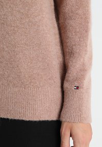 Roze gebreide trui met een zachte textuur, geribde zoom en een klein geborduurd logo in rood, wit en blauw op de manchet.