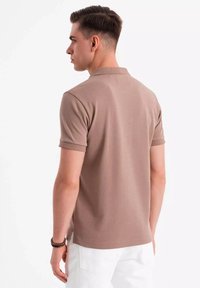 Camisa polo de algodón marrón con mangas cortas, que presenta un cuello clásico y sutiles detalles de costura, exhibida en un modelo desde un ángulo trasero.
