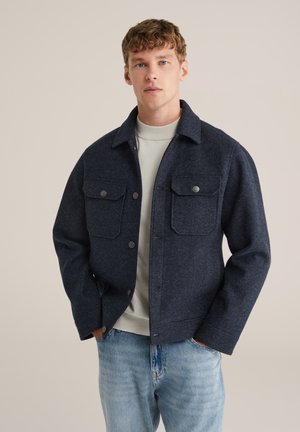 Giacca in misto lana blu navy con colletto, due tasche sul petto e chiusura a bottoni, indossata sopra un maglione grigio chiaro.