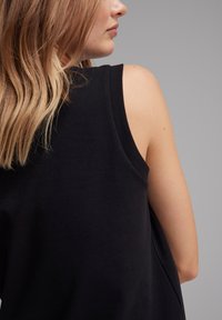Top nero senza maniche, con una vestibilità rilassata, realizzato in cotone, con scollatura rotonda e texture liscia. Mostra parte della spalla e dei capelli.