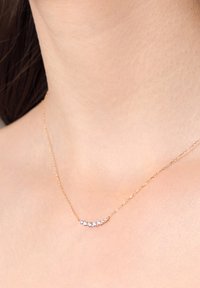 Złoty naszyjnik z łańcuszkiem, wyposażony w zakrzywioną belkę z pięcioma małymi, okrągłymi przezroczystymi kryształami, na tle gładkiej skóry.