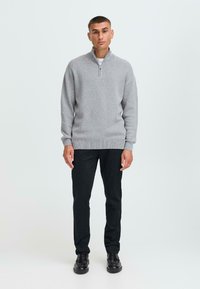 Grijze gebreide trui met een halve rits kraag, ribbelzoom, gedragen over een witte shirt en gecombineerd met zwarte broek en zwarte schoenen.