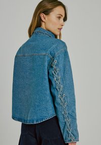Gaišā zila džinsu jaka ar brīvu piegriezumu, ar mežģīņu detaļām gar piedurknēm un klasisku apkakli.
