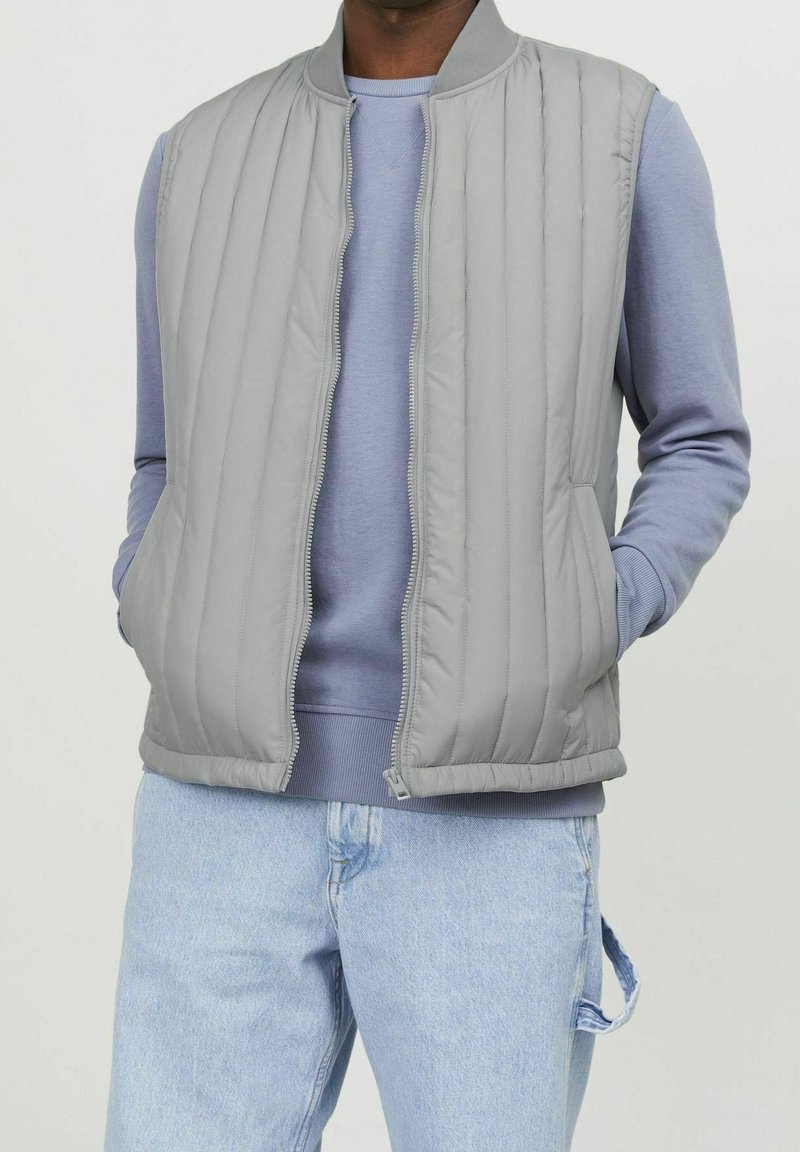 Grå kviltad väst med rund halsringning, med framfickor och dragkedjestängning, lagd över en ljusblå sweatshirt. Denimbyxor nedanför.