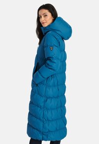 Lang pufferjas in felblauw met een capuchon, voorzien van een gestikte uitstraling, ritszakken en een logo badge op de linkerarm.