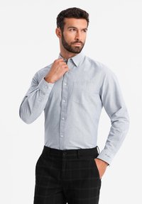 Camisa de botón azul claro, hecha de una tela con textura suave, que presenta un bolsillo en el pecho y botones en contraste, combinada con pantalones de cuadros negros.