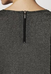 Pull noir en texture métallique avec un col haut et une fermeture éclair courte au dos avec des détails en métal argenté.
