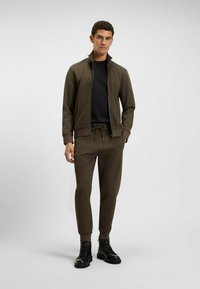Zielono-oliwkowy dres z kurtką na zamek i spodniami z troczkami; gładki materiał; dopasowany krój; zestawiony z czarnymi butami na sznurowadła.