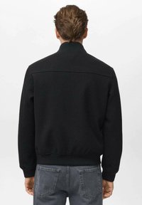 Chaqueta bomber negra con un acabado texturizado, cuello y puños acanalados. Presenta un diseño limpio en la parte posterior, combinada con jeans de mezclilla grises.