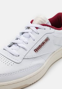 Weiße Ledersneaker mit roten Akzenten, ausgestattet mit einer strukturierten Sohle, einem durchlässigen Zehenbereich und einem Markenetikett auf der Zunge.