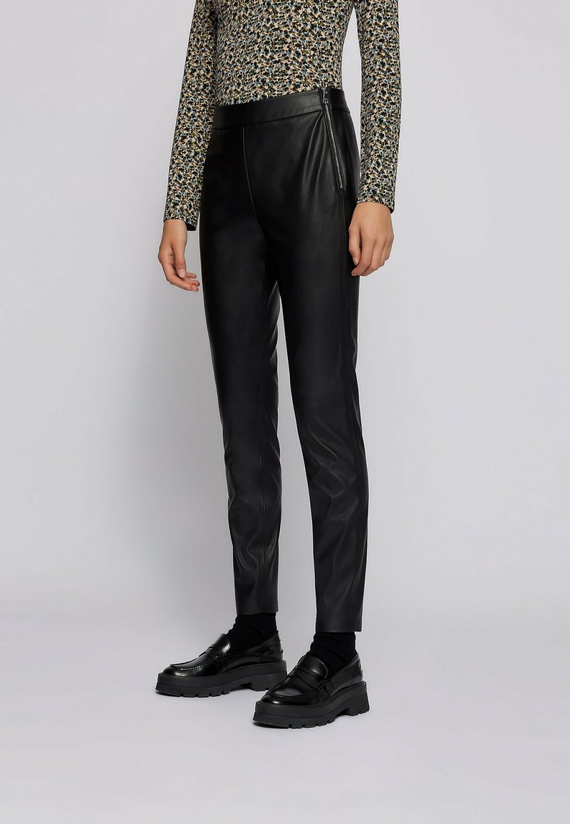Femme portant un pantalon en cuir noir slim, des mocassins noirs épais, des chaussettes noires et un haut à manches longues à motifs sur un fond gris uni.